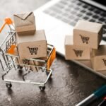 selective focus of toy shopping cart with small carton boxes near laptop, e-commerce concept